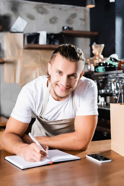 Happy barista looking at camera while writing in notebook near smartphone with blank screen on bar counter — Stock Photo