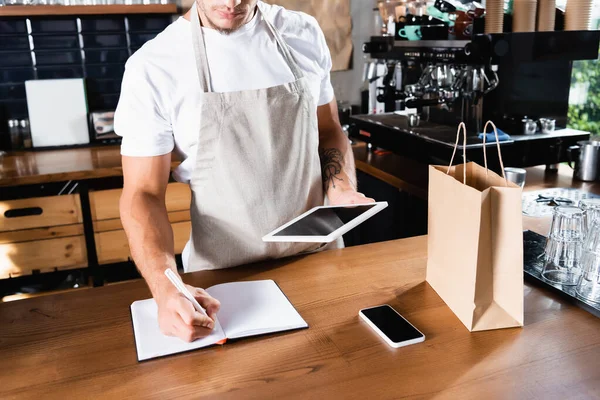 Vista recortada de barista en delantal escrito en portátil mientras sostiene la tableta digital cerca de teléfono inteligente y bolsa de papel - foto de stock