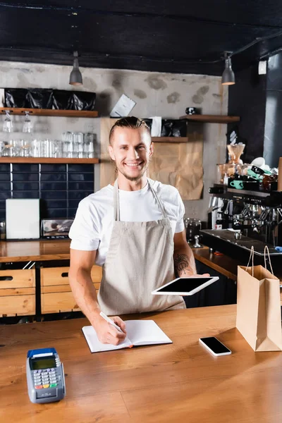 Bello, giovane barista sorridente alla macchina fotografica tenendo tablet digitale e scrivendo nel taccuino al bancone del bar — Foto stock