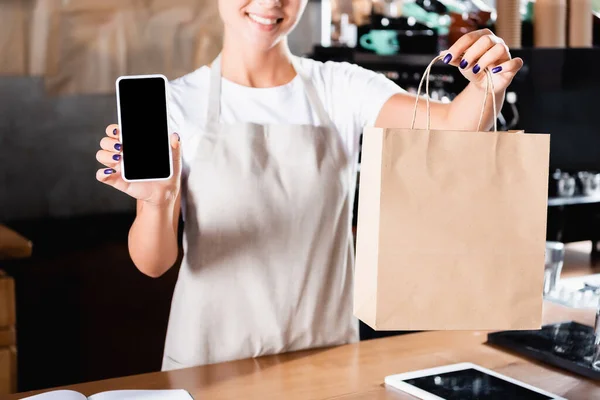 Vista recortada de barista sosteniendo bolsa de papel y mostrando teléfono inteligente con pantalla en blanco, fondo borroso - foto de stock