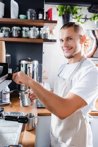 Joven sonriente barista mirando a la cámara mientras sostiene la taza de leche metálica cerca del vapor de la máquina de café - foto de stock