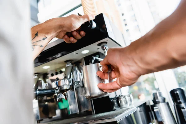 Vue partielle du barista tenant une tasse de lait métallique près d'un vapeur de machine à café au premier plan flou — Photo de stock