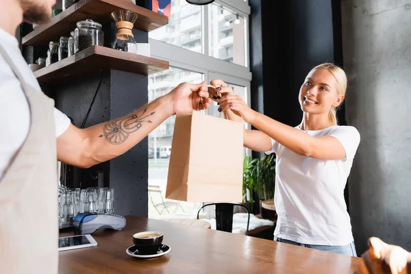 Donna bionda sorridente che prende la borsa di carta dal barista tatuato su primo piano sfocato — Foto stock