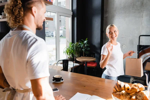 Cheerful blonde woman holding paper bag and waiving hand to barista on blurred foreground — Stock Photo