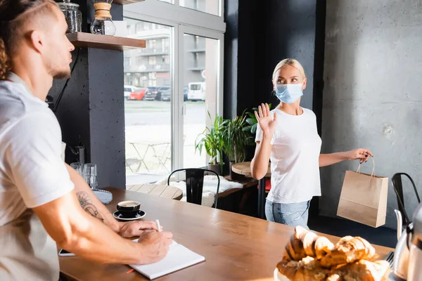 Joven mujer en máscara médica sosteniendo bolsa de papel y agitando la mano a barista en primer plano borrosa - foto de stock