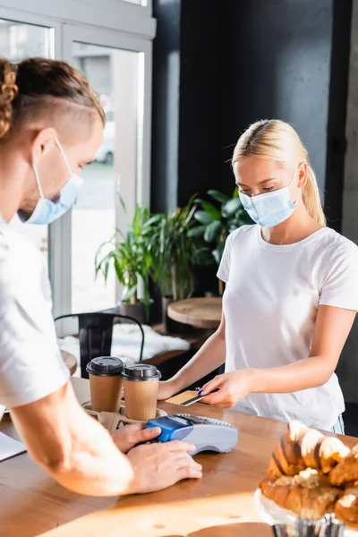 Woman in medical mask paying for coffee to go through payment terminal near barista on blurred foreground — Stock Photo