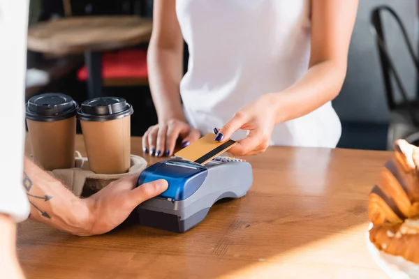 Vista parcial de la mujer en posesión de tarjeta de crédito cerca de barista con terminal de pago en primer plano borroso — Stock Photo