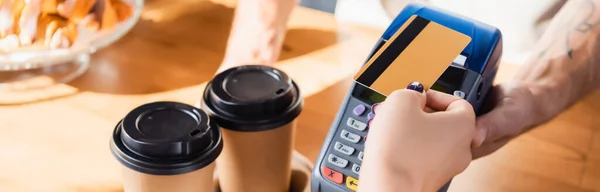 Vista recortada de la mujer que paga con tarjeta de crédito a través de la terminal cerca de café para llevar en la cafetería, pancarta - foto de stock