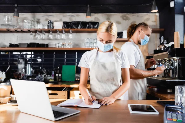 Giovane cameriere che scrive in quaderno vicino a gadget e barista che lavora sullo sfondo — Foto stock