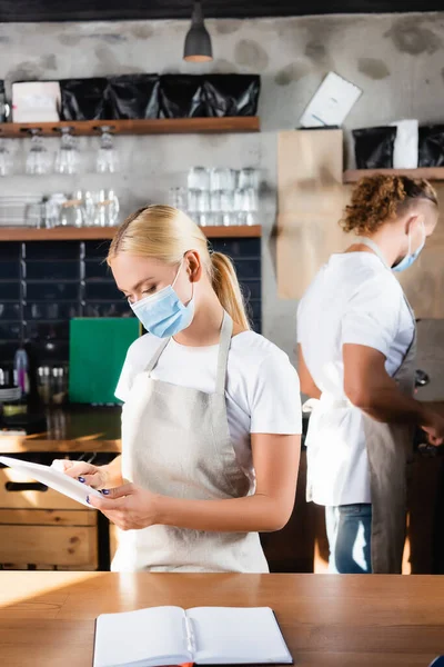 Giovane barista bionda in maschera medica utilizzando tablet digitale vicino collega su sfondo sfocato — Foto stock