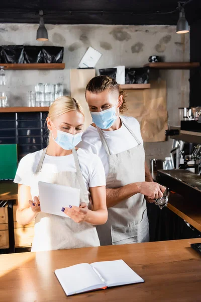 Young waiter in medical mask holding digital tablet near barista with portafilter and blank notebook on bar counter — Stock Photo