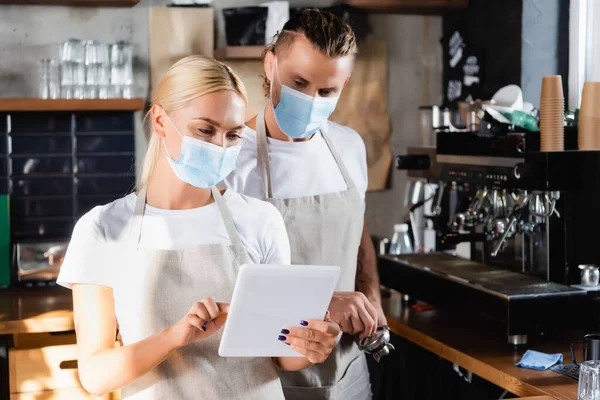 Junge blonde Kellnerin in medizinischer Maske mit digitalem Tablet in der Nähe von Barista mit Portafilter — Stockfoto