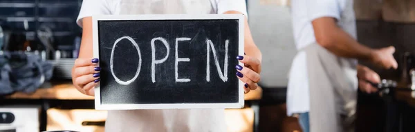 Cropped view of barista holding board with open lettering near colleague on blurred background, banner — Stock Photo