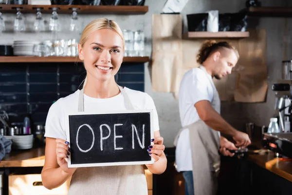 Young smiling waiter holding board with open lettering near barista working on blurred background — Stock Photo