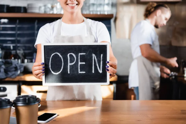 Cropped view of smiling waiter holding board with open lettering near barista working on blurred background — Stock Photo
