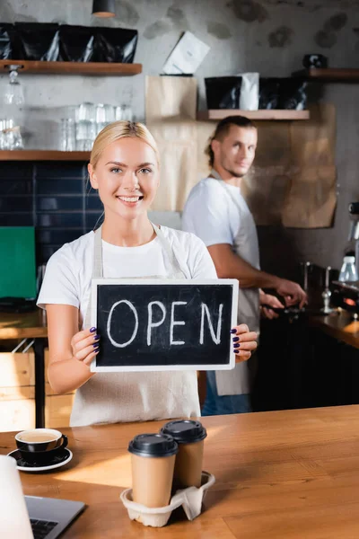 Bionda, barista sorridente che tiene la tavola con lettere aperte vicino al collega su sfondo sfocato — Foto stock