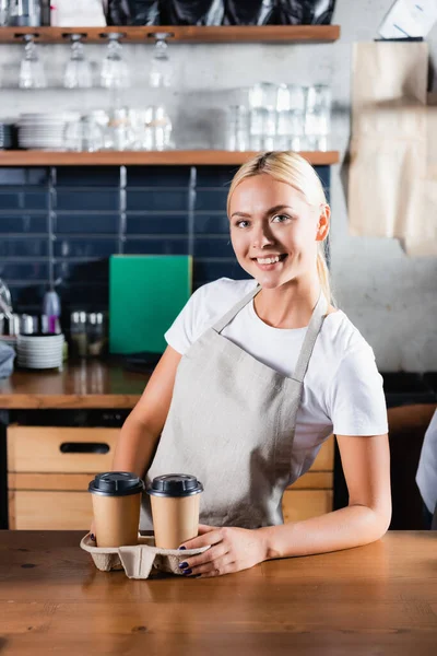 Alegre rubia barista mirando la cámara en bar mostrador cerca de café para ir - foto de stock