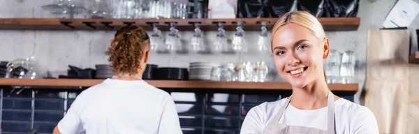 Joven rubia barista sonriendo en cámara cerca colega en fondo, pancarta - foto de stock