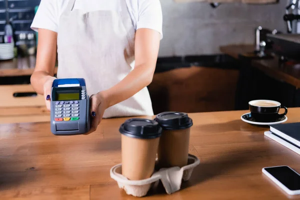 Vista recortada del camarero en la terminal de pagos de la tenencia del delantal cerca del café para ir en primer plano borroso — Stock Photo