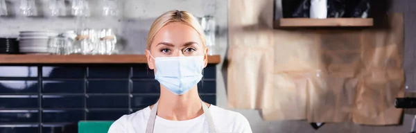 Young blonde barista in medical mask looking at camera, banner — Stock Photo