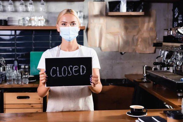 Barista loira em máscara médica segurando bordo com letras fechadas perto bar contador — Fotografia de Stock
