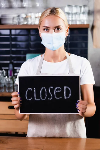 Blonde barista in medical mask holding board with closed lettering in cafe — Stock Photo