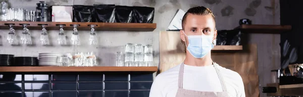 Barista in maschera medica guardando la fotocamera mentre in piedi sul posto di lavoro in caffè, banner — Foto stock