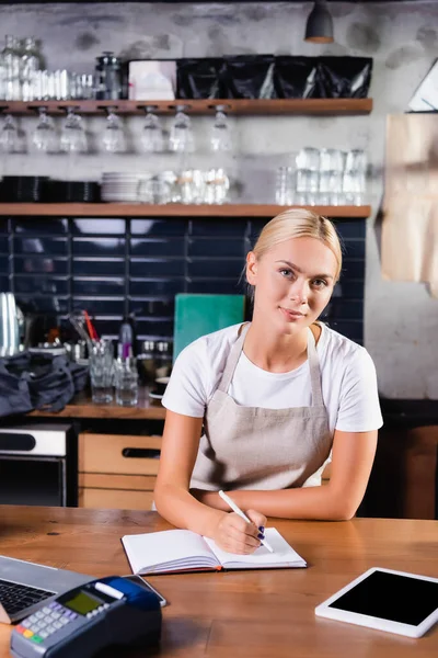 Young blonde barista writing in notebook near digital tablet and payment terminal on blurred foreground — Stock Photo