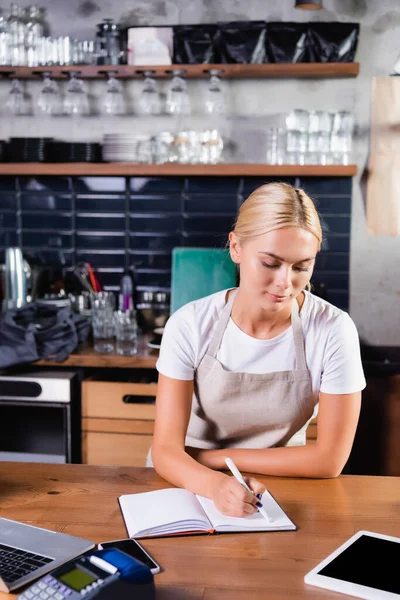 Young blonde barista writing in notebook near gadgets and payment terminal on blurred foreground — Stock Photo