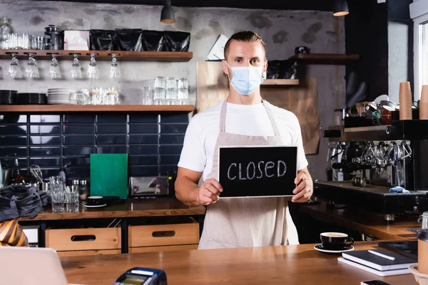 Young barista in medical mask holding board with closed lettering at workplace in coffee house — Stock Photo