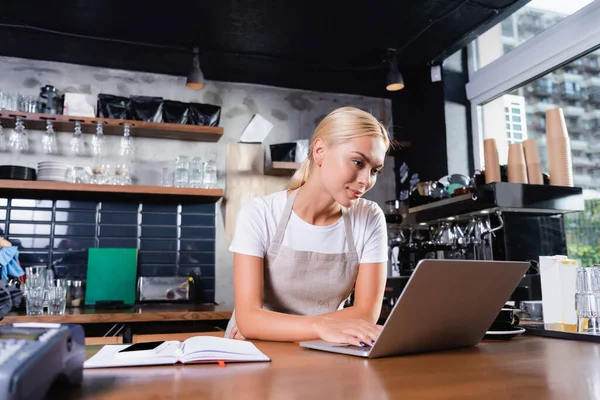 Jovem barista loira usando laptop perto notebook no balcão de bar, foreground borrado — Fotografia de Stock