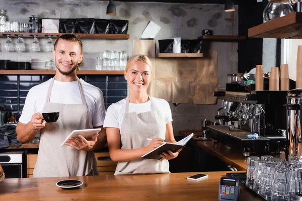 Giovani baristi in grembiuli sorridenti alla macchina fotografica mentre in piedi al bancone del bar — Foto stock