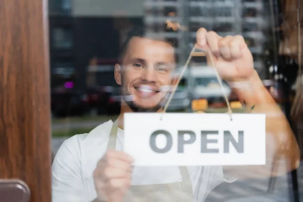 Smiling barista hanging card with open lettering on entrance of coffee house — Stock Photo