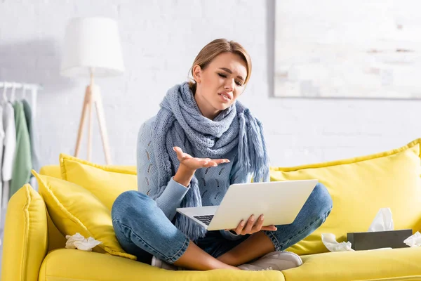 Femme mécontente et malade utilisant un ordinateur portable tout en étant assise près des serviettes sur le canapé — Photo de stock