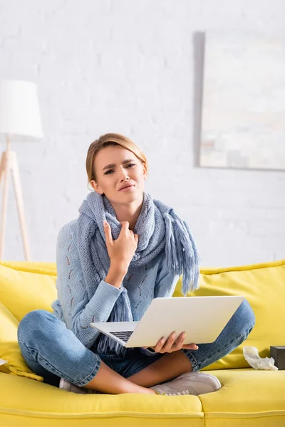 Sick and disappointed woman in scarf holding laptop on couch — Stock Photo