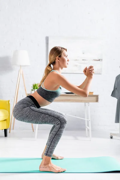 Vue latérale de la jeune sportive faisant squat sur tapis de fitness à la maison — Photo de stock