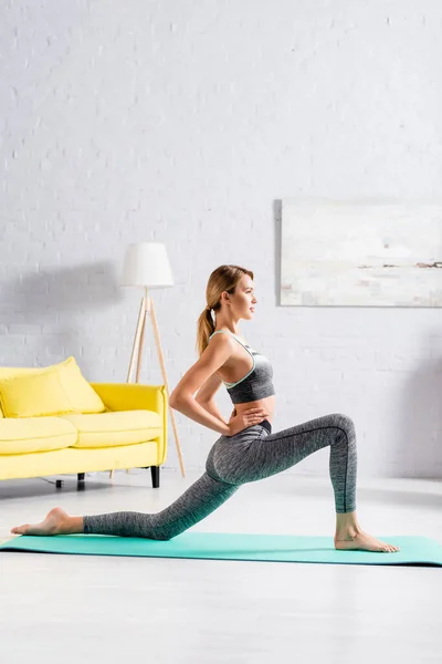 Vista laterale della sportiva bionda in piedi in posa yoga sul tappeto fitness a casa — Foto stock