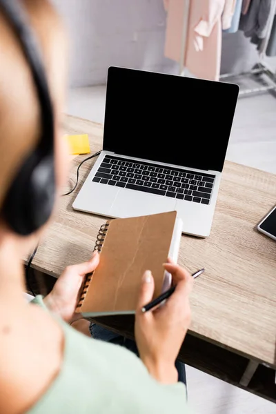 Laptop with blank screen on table near young woman in headset holding notebook on blurred foreground — Stock Photo