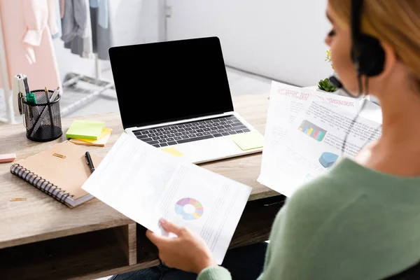 Portátil con pantalla en blanco en la mesa cerca de freelancer en auriculares con papeles con gráficos en primer plano borroso - foto de stock