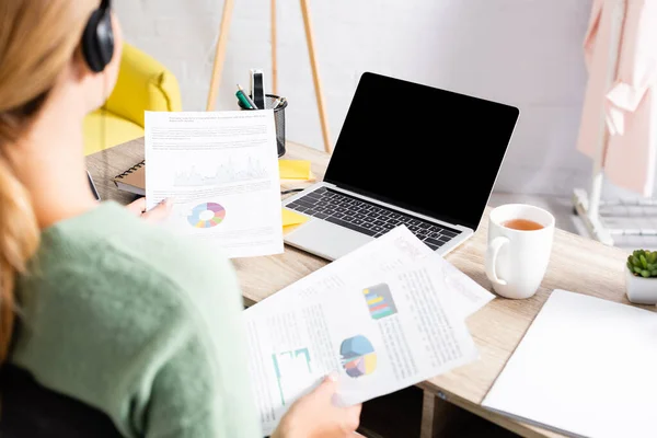 Laptop with blank screen near cup of tea and freelancer in headset holding documents on blurred foreground — Stock Photo