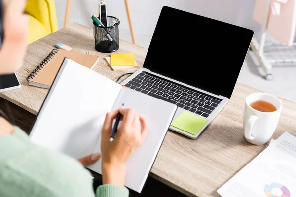 Laptop with blank screen near cup of tea and papers on table near freelancer writing on notebook on blurred foreground — Stock Photo