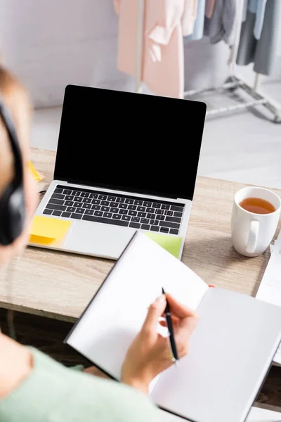 Laptop with blank screen near cup of tea and freelancer in headset writing on notebook on blurred foreground — Stock Photo