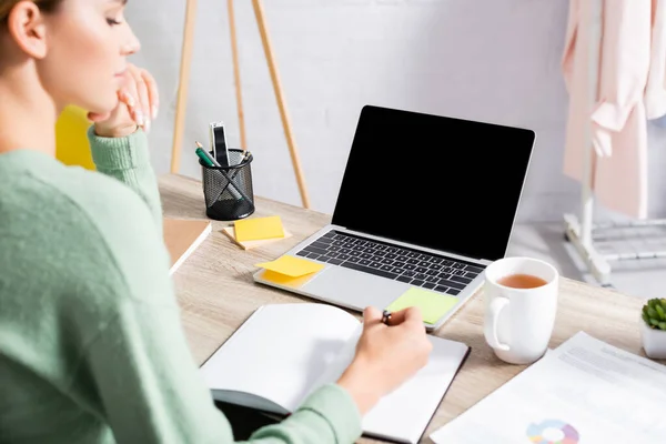 Laptop mit leerem Bildschirm bei Tasse Tee und Freiberufler schreiben auf Notizbuch und Papiere auf verschwommenem Vordergrund — Stockfoto