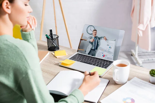 Geschäftsmann zeigt mit Finger in der Nähe von Flipchart auf Bildschirm von Laptop in der Nähe von Freelancer Schreiben auf Notizbuch während Webinar auf unscharfen Vordergrund — Stockfoto