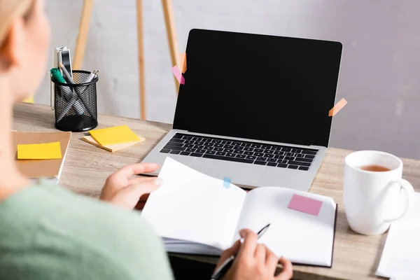 Laptop com tela em branco perto de notas pegajosas, xícara de chá e freelancer segurando caneta e notebook em primeiro plano desfocado — Fotografia de Stock