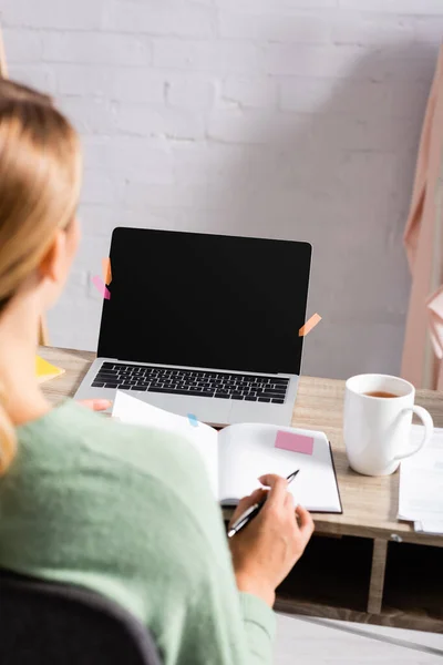 Portátil con notas adhesivas y pantalla en blanco cerca de taza de té y freelancer con portátil y pluma en primer plano borroso - foto de stock