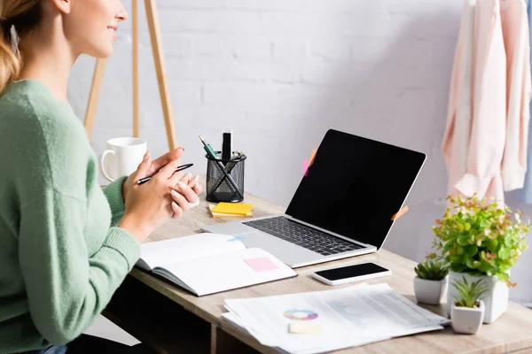 Vista recortada de freelancer sorrindo segurando caneta perto de dispositivos com tela em branco e papéis em primeiro plano desfocado em casa — Fotografia de Stock