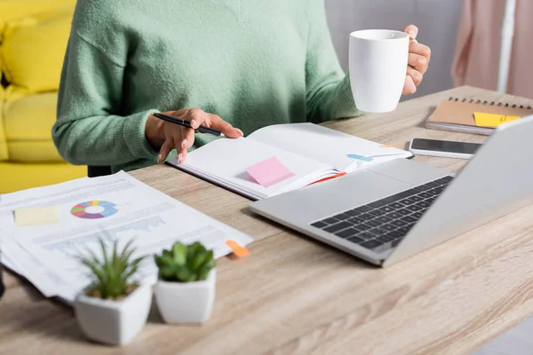 Vista cortada do teleworker que prende o copo e a pena perto do caderno, dos papéis e dos dispositivos no primeiro plano borrado — Fotografia de Stock