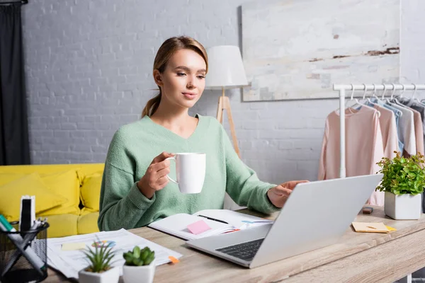 Freelancer con taza usando laptop cerca de notebook y documentos en primer plano borroso en casa - foto de stock