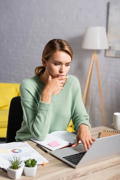 Pensive freelancer using laptop while working near notebook and documents on blurred foreground — Stock Photo